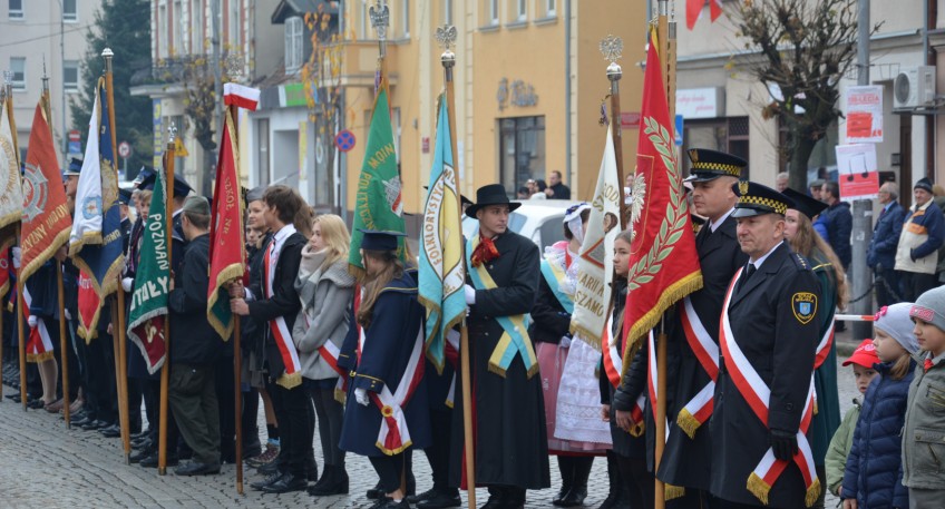 Grupa ludzi w różnym wieku ustawiona w jeden rząd. W grupie tych osób znajdują się przedstawiciele różnych jednostek i organizacji ubranych w strój galowy. Niektórzy przedstawiciele trzymają swój sztandar, a takżeubrani są we wstęge.