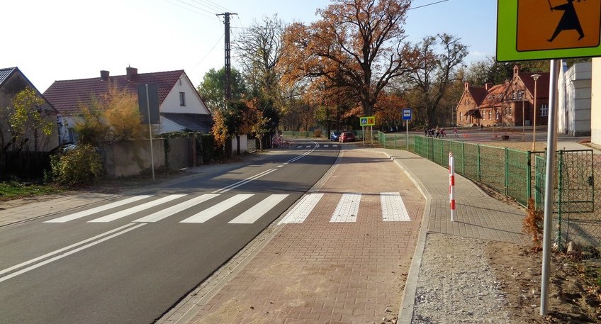 Ulica  przejściem dla pieszych. Z lewej strony są zabudowania, po prawej zatoczka autobusowa oraz chodnik dla pieszych.