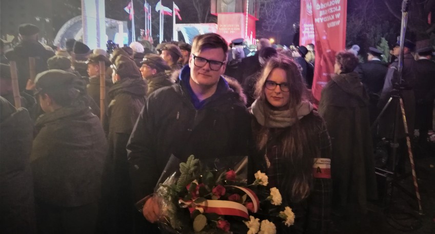 Para uczestników poznańskich obchodach setnej rocznicy wybuchu Powstania Wielkopolskiego. Mężczyzna trzyma kwiaty z biało-czerwoną wstęgą. Kobieta na ramieniu ma opaskę AK. W tle inni uczestnicy. 