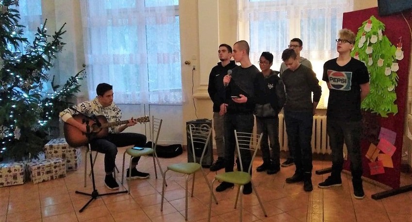 Grupa młodzieży, od lewej siedzi chłopak trzymający gitarę, następnie stoi sześć chłopaków w różnym wzroście. Jeden z nich trzyma mikrofon.