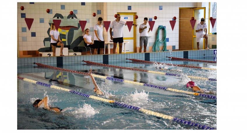 W basenie na wyznaczonych torach pływają zawodnicy. Na zewnętrznej stornie basenu stoją sędziowie oraz osoby mierzące czas.