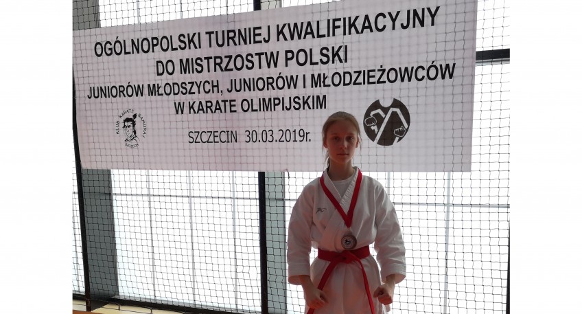 Karolina Kukawka ubrana w biały strój karate przepasany czerwonym pasem. Na szyi wisi brązowy medal na czerwonej wstędze. W tle widać baner z napisem: 