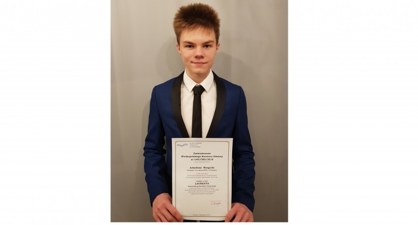 Arkadiusz Wargacki trzymające dyplom oświadczający, że został Laureatem Wojewódzkiego Konkursu Chemicznego. Chłopak ubrany jest w garnitur.