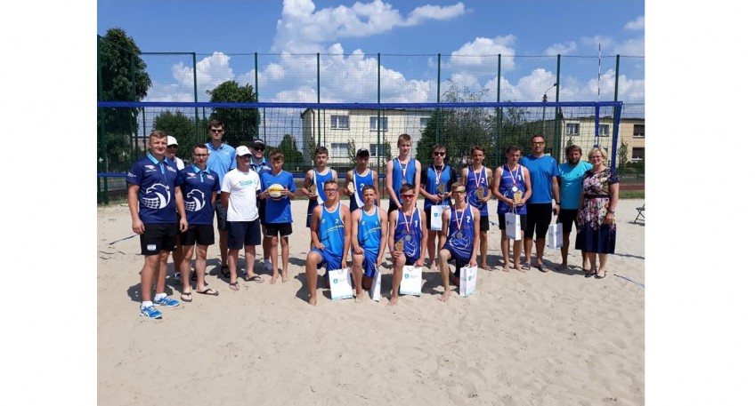 Grupa uczestników zawodow sportowych. Część z nich trzyma upominki. Zdjęcie jest zrobione na tle boiska plażowego do piłki siatkowej