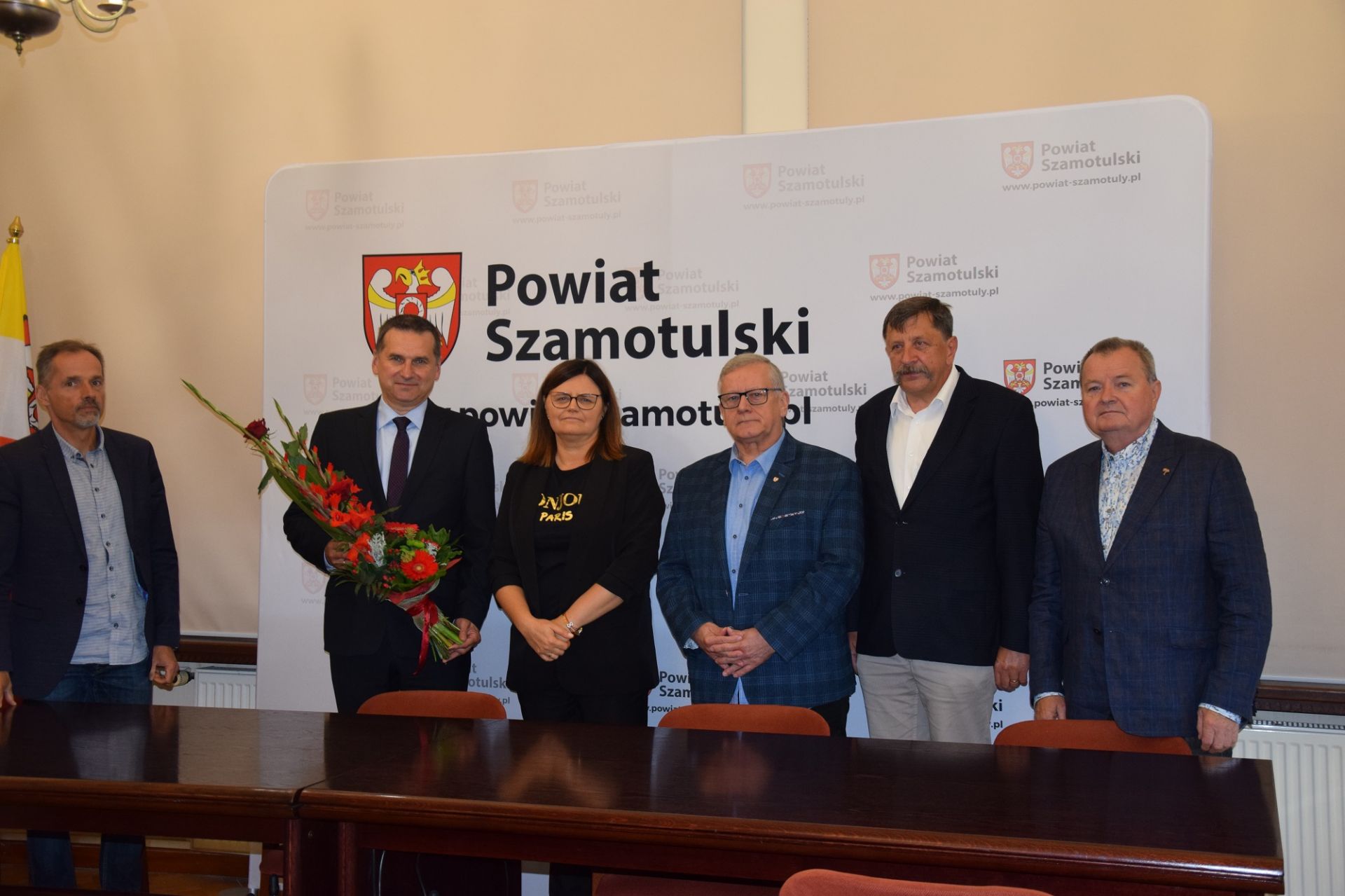zdjęcie grupowe na tle planszy z herbem powiatu szamotulskiego. Na zdjęciu ustawieni są kolejno, od lewej: Wicestarosta Szamotulski Pan Rafał Zimny, Dyrektor SP ZOZ w Szamotułach Pan Remigiusz Pawelczak, Starosta Szamotulski Pani Beata Hanyżak, członek zarządu Pan Kazimierz Michalak, Pan Andrzej Grzeszczyk oraz Pan Maciej Trąbczyński