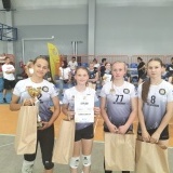 Siatkarki w szarych koszulkach, ubrane na sportowo, z nagrodami, dyplomami i pucharem, w tle trybuny, kibice i inne zawodniczki