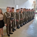 Około 20 uczniów ustawionych w szeregu w zielonych mundurach wraz z dowódcą i nauczycielką na sali