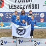 Zawodnicy LUKS „Gry Logiczne” z Wronek na podium na tle banerów imprezy, przed sobą trzymają logo klubu 